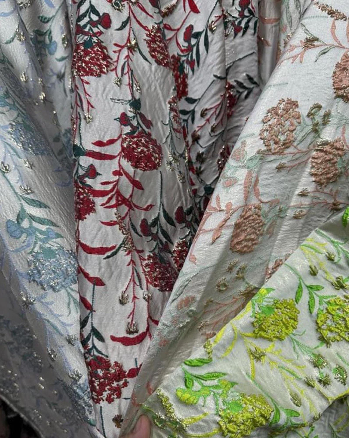 May include: A textured off-white fabric featuring an intricate floral pattern with embroidered details in shades of red, green, blue, and yellow. The material has a subtle sheen and includes metallic accents woven throughout the botanical design.